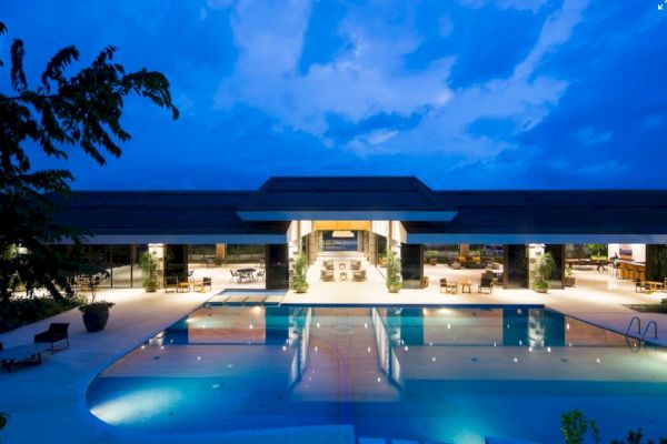 The image displays a well-lit, modern building with a large swimming pool in front, taken at dusk with a blue sky backdrop.