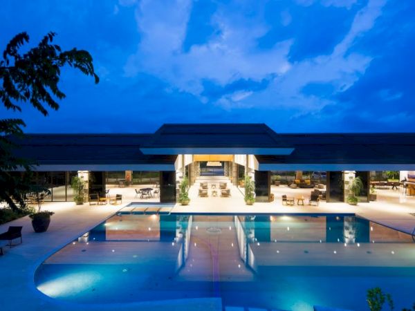 The image displays a well-lit, modern building with a large swimming pool in front, taken at dusk with a blue sky backdrop.