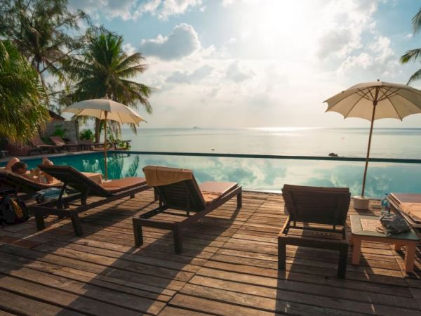 A serene beachside setting with lounge chairs, umbrellas, and a wooden deck overlooking a calm sea and a view of the sunset or sunrise.