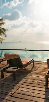 A serene poolside scene with tanning beds, umbrellas, and palm trees overlooking a calm ocean under a partly cloudy sky and distant horizon.