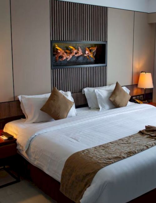 A well-lit hotel room with a king-sized bed, two bedside tables with lamps, cushions, and a decorative towel arrangement on the bed.