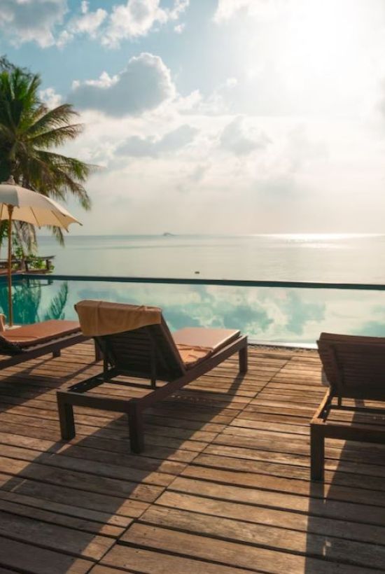 Wooden deck with sun loungers, umbrellas, and a swimming pool, overlooking a scenic ocean view under a partly cloudy sky.