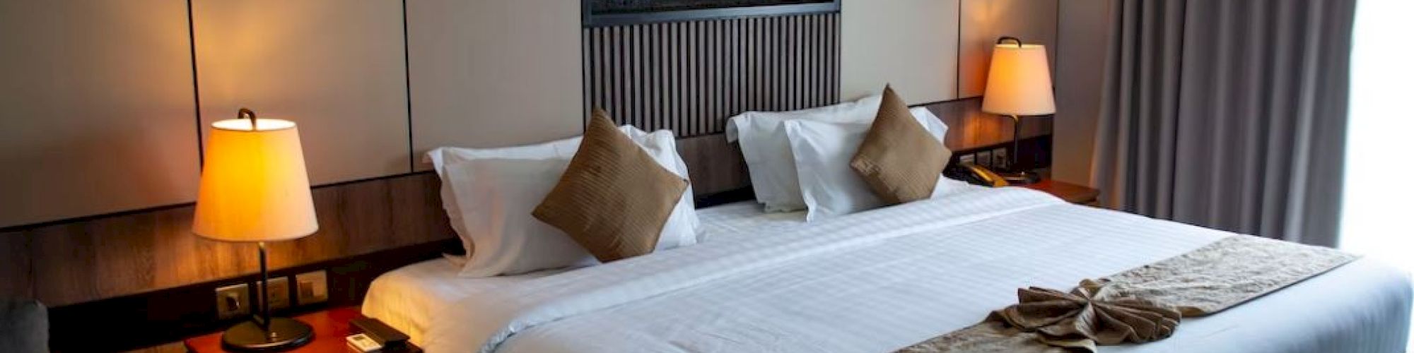 A cozy bedroom with a large bed, two bedside tables with lamps, pillows, and a decorative piece above the bed, all in soft, warm lighting.