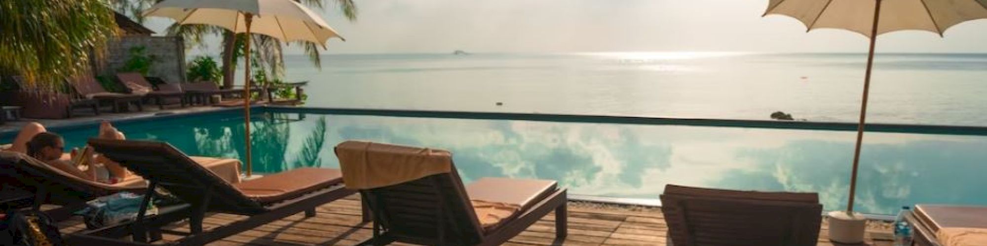 A serene poolside setting with lounge chairs, umbrellas, palm trees, and a stunning view of the ocean under a partly cloudy sky during daytime, ending the sentence.