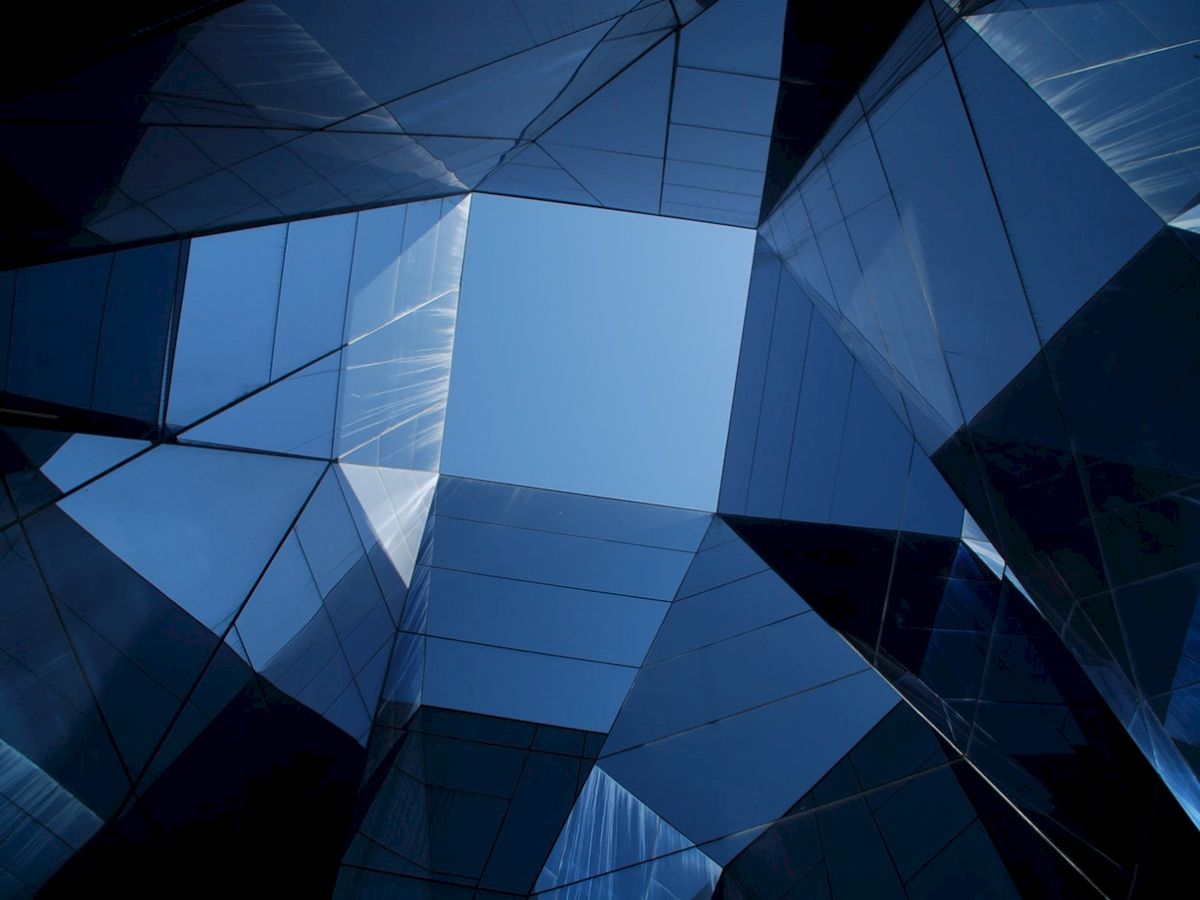 The image shows a geometric architectural structure made of reflective glass panels, with a view of the blue sky in the center.