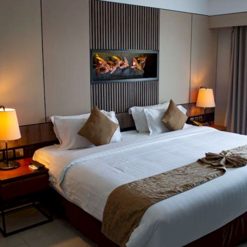A modern hotel room features a large bed, two bedside lamps, a decorative wall feature, and a small round table by the window with curtains.
