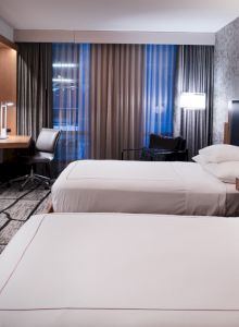 A modern hotel room with two double beds, a wall-mounted TV, a desk with a chair, lamps, and curtains on windows, creating a cozy atmosphere.