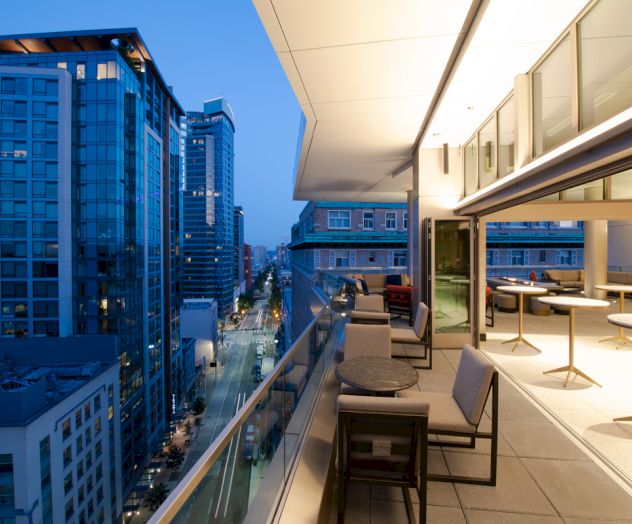 A modern, city balcony with seating areas and tables, overlooking a busy urban street lined with tall buildings.