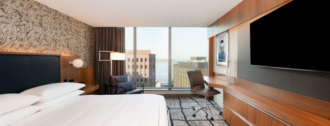A modern hotel room with a large bed, desk, chair, flatscreen TV, city view from the window, contemporary decor, and patterned carpeting.