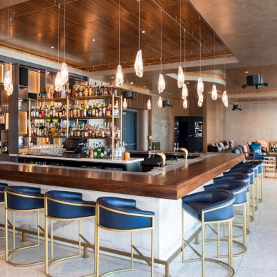 The image shows a modern bar with blue stools, a well-stocked bar, hanging lights, and a seating area in the background.