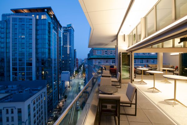 A modern cityscape scene in the evening with tall buildings, a busy street below, and an elegant balcony with seating and tables overlooking the view.