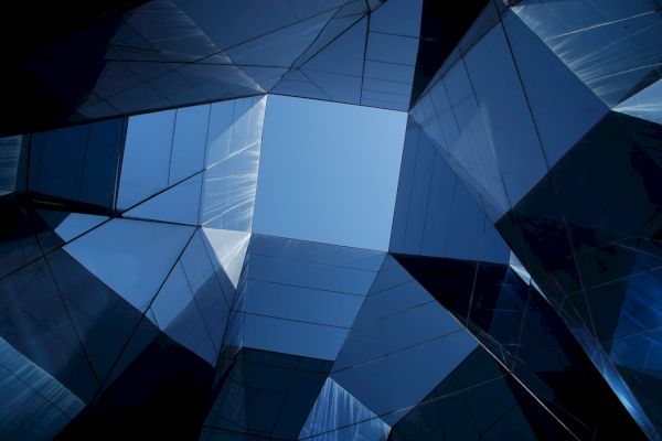 The image shows a geometric architectural structure made of reflective glass panels, forming a polygonal opening that reveals a clear blue sky.