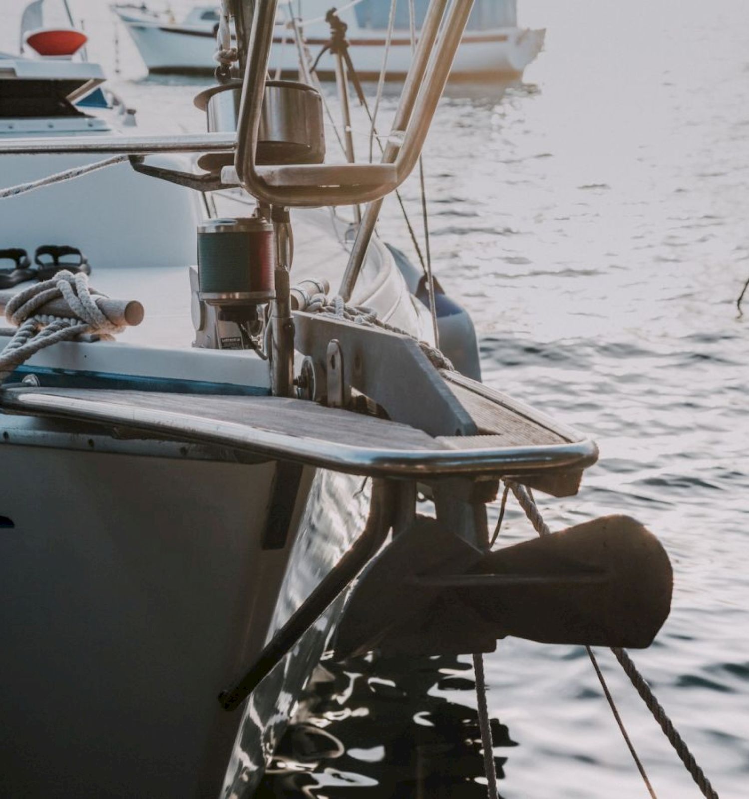 The image shows the bow of a sailboat anchored in a calm marina, with ropes and an anchor visible, next to other boats.