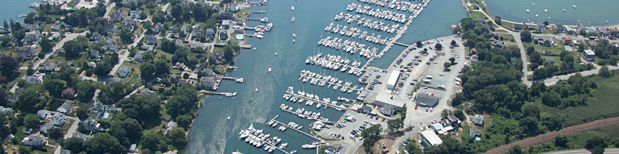 Aerial view of a marina with numerous docked boats, nearby residential areas, green spaces, and a stretch of railway tracks in the foreground.