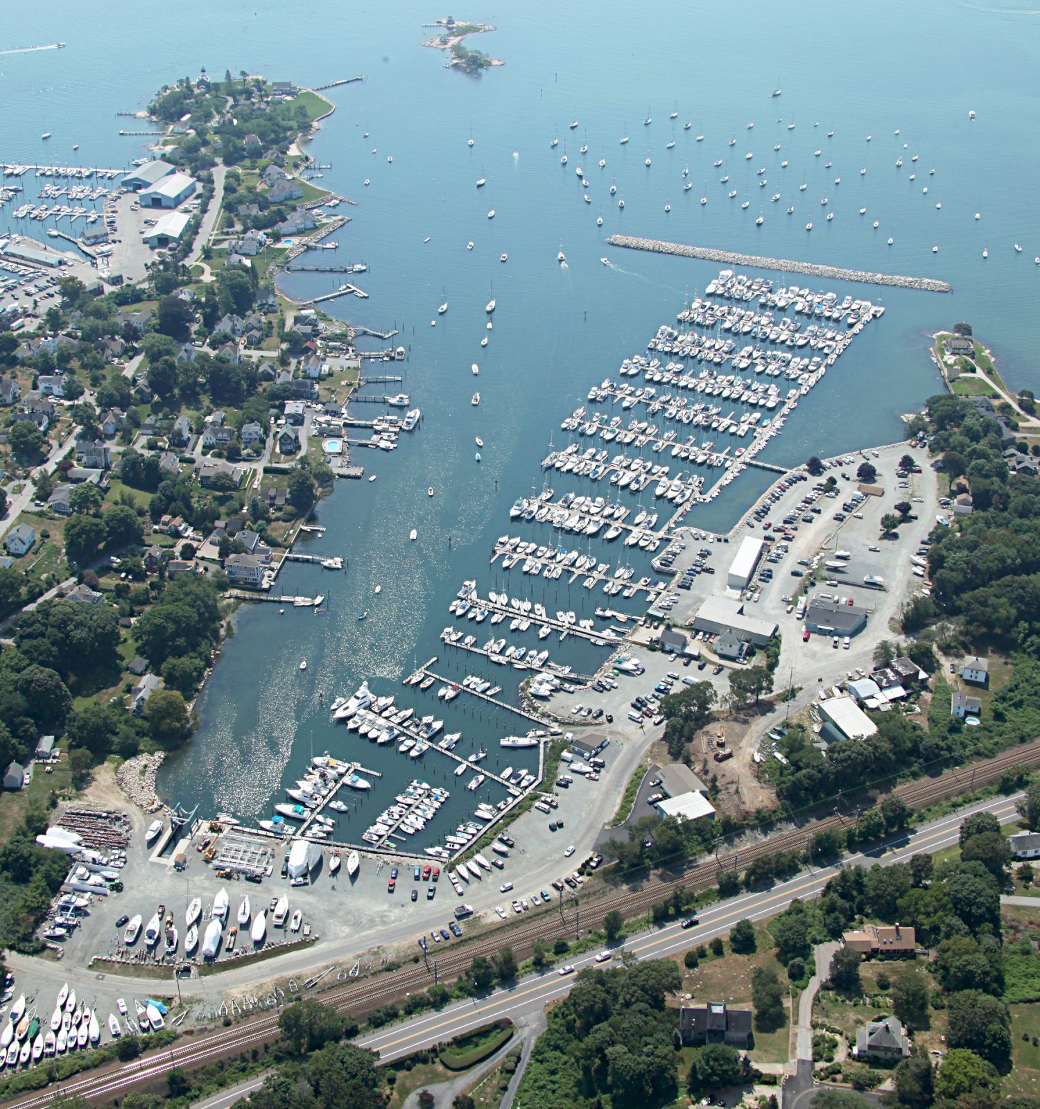 Aerial view of a coastal town with numerous boats docked in a marina, surrounded by lush greenery, residential areas, and railway tracks.