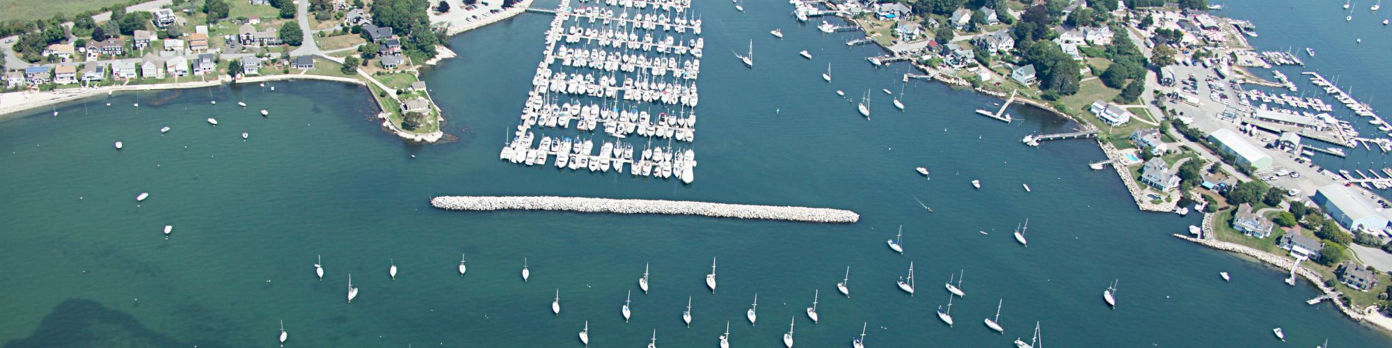 An aerial view of a marina with numerous docked boats and sailboats in the water, surrounded by a coastal town and greenery, ending the sentence.