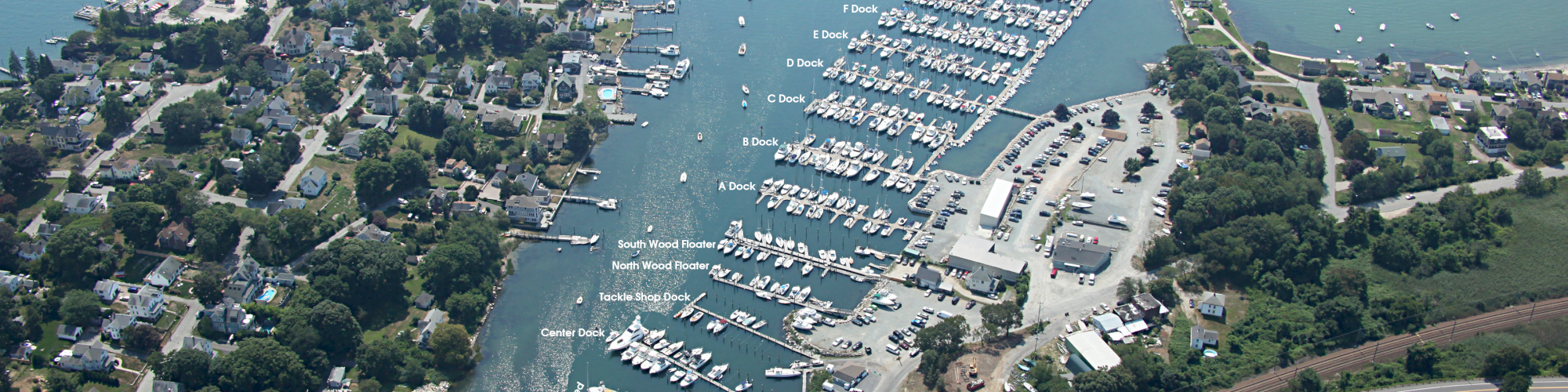 An aerial view of a coastal area featuring a marina with numerous boats, surrounding residential areas, and a railway line in the foreground.