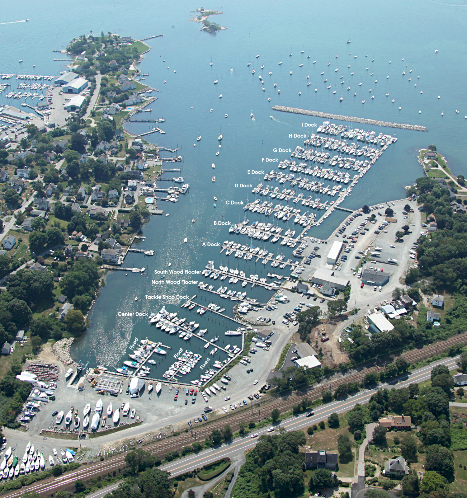 An aerial view of a coastal marina with numerous boats docked, surrounded by residential areas, water, and a visible railway line.