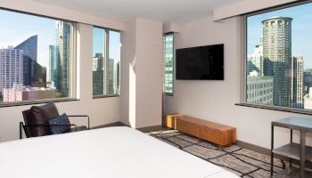 A modern bedroom with large windows showcasing a cityscape, a wall-mounted TV, an armchair, and a low wooden console.