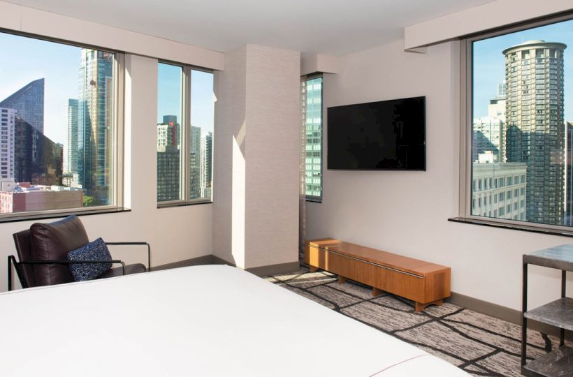 A modern bedroom with large windows showcasing a cityscape, a wall-mounted TV, an armchair, and a low wooden console.