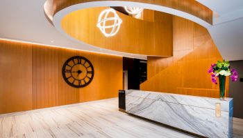 A modern lobby with a wooden spiral staircase, marble reception desk, wall clock, and a flower arrangement is shown in the image.