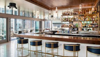 This image shows a modern bar with a long counter, blue stools, hanging lights, and a wide array of bottled drinks on shelves. The sentence ends.