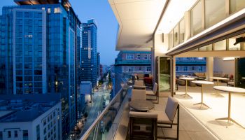 A cityscape at dusk with modern high-rise buildings, viewed from a balcony with outdoor seating and tables, extending from an indoor space.