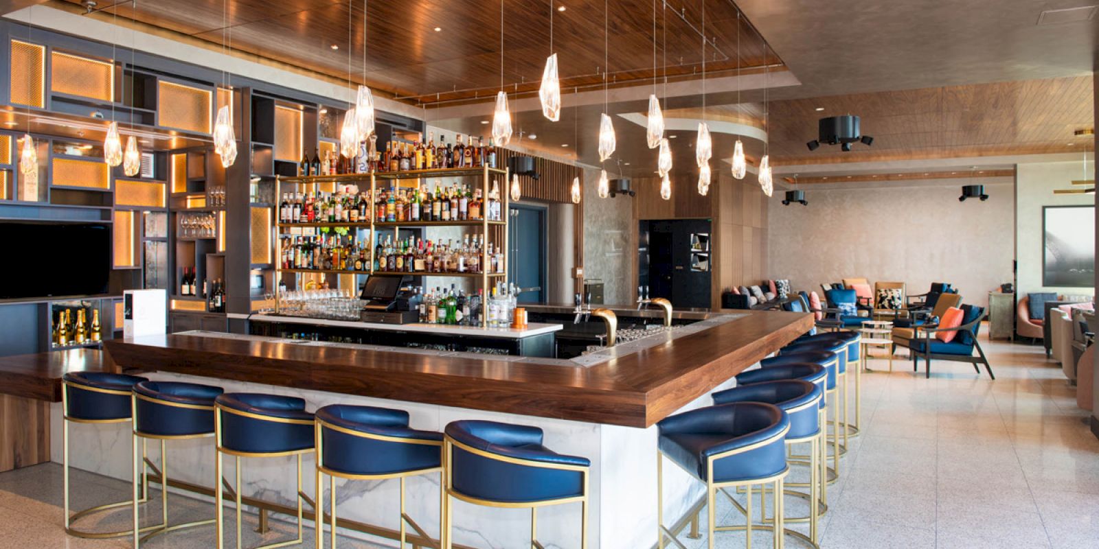 This image shows an upscale bar interior with a central bar counter, blue and gold barstools, hanging lights, and a wall of liquor bottles.