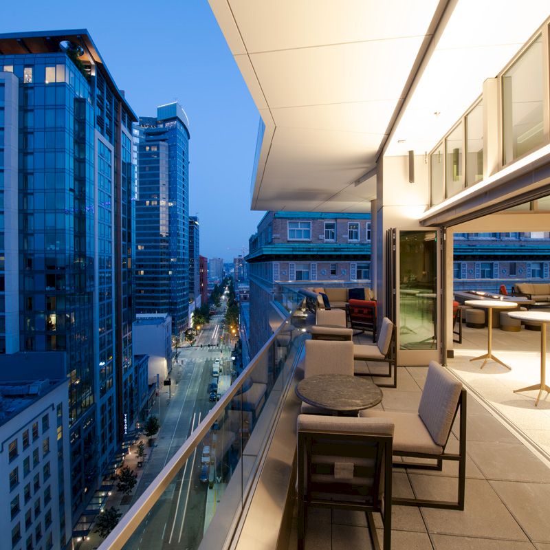 A modern cityscape at dusk featuring a stylish outdoor balcony area with tables, chairs, glass railings, and tall buildings in the background.