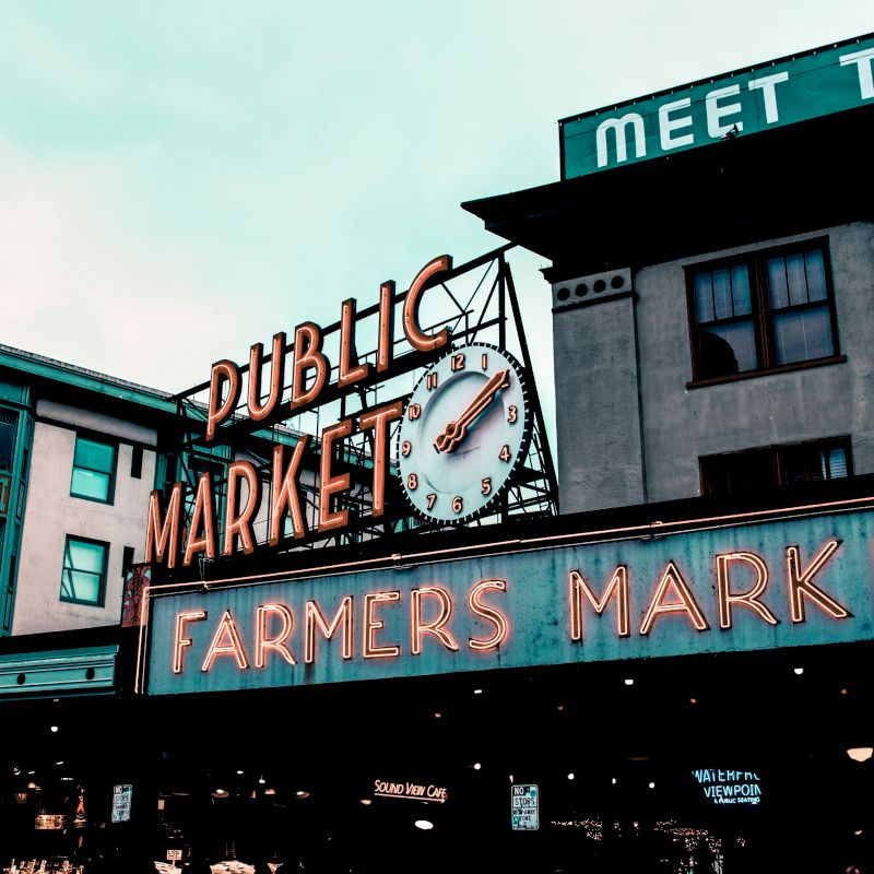 Image shows the exterior of a public farmers market with neon signs and a clock, surrounded by buildings and an overcast sky.