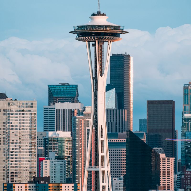 The image shows the Space Needle with the Seattle skyline in the background. The iconic tower stands tall among surrounding modern buildings.