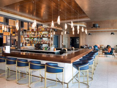 A modern bar with a wooden counter, bar stools, pendant lights, and a well-stocked liquor shelf, located in an upscale lounge area.