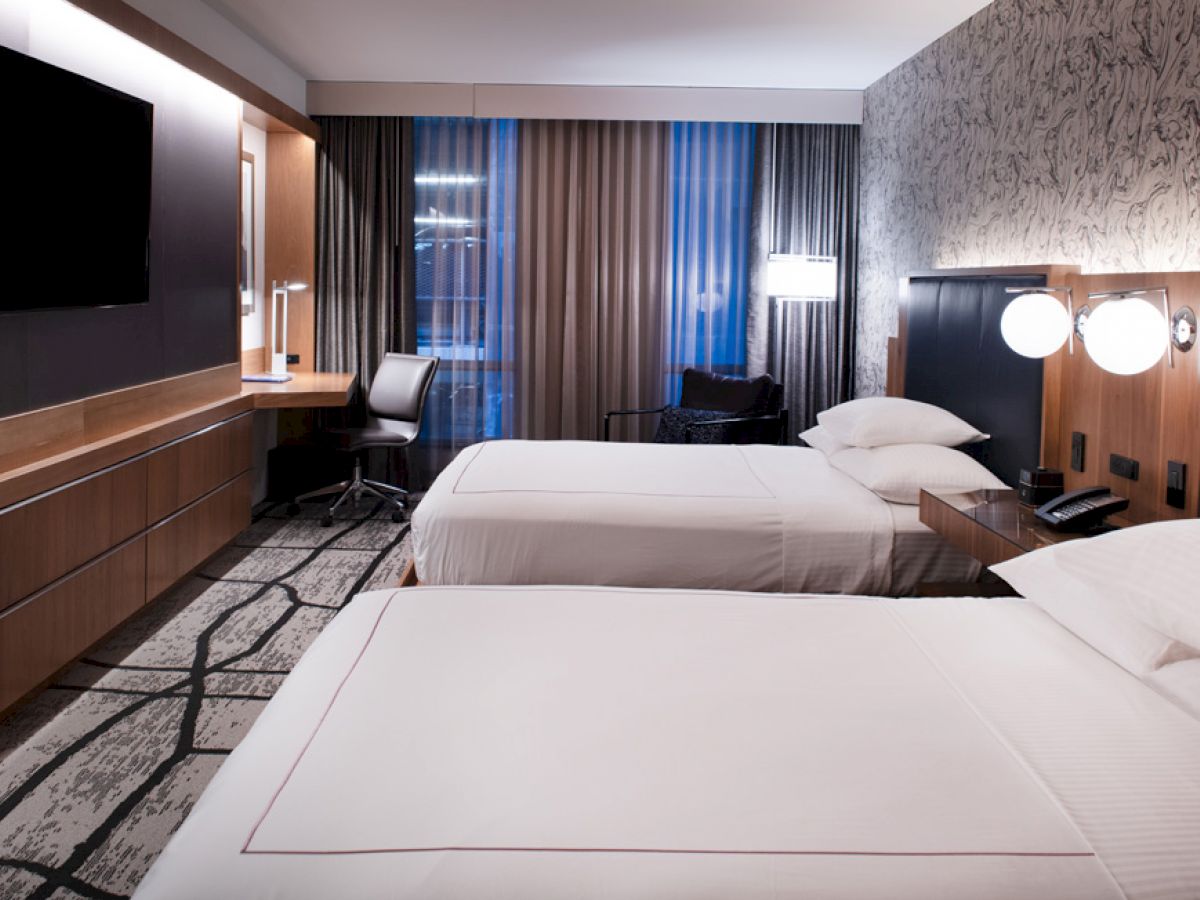 A modern hotel room with two neatly-made beds, a large TV, a desk with a chair, lamps on the bedside tables, and patterned wallpaper.