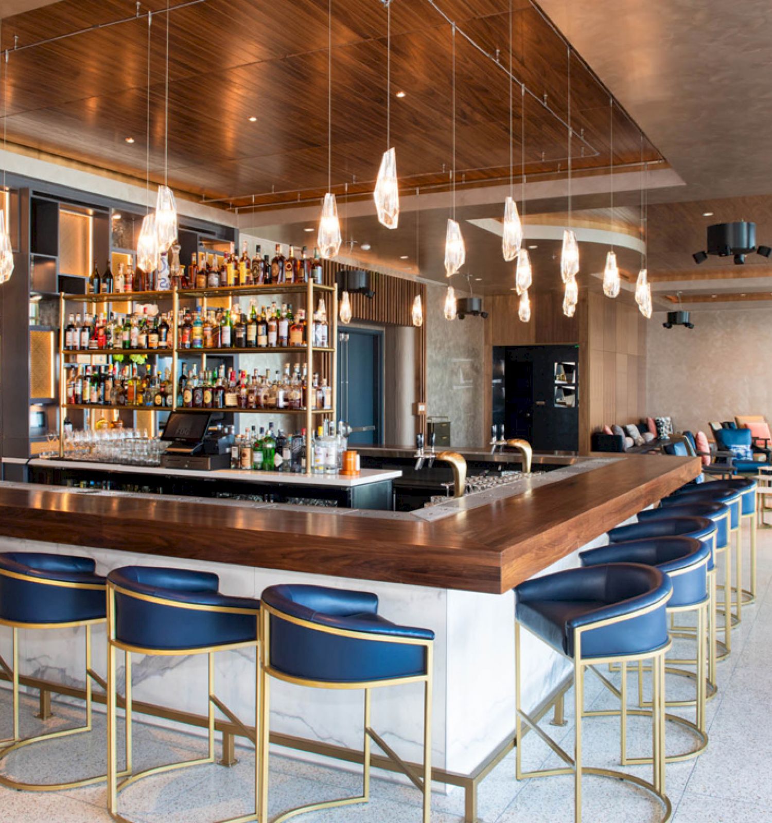 A modern bar with blue cushioned bar stools, hanging pendant lights, a well-stocked back bar, and additional seating with tables and chairs in the background.