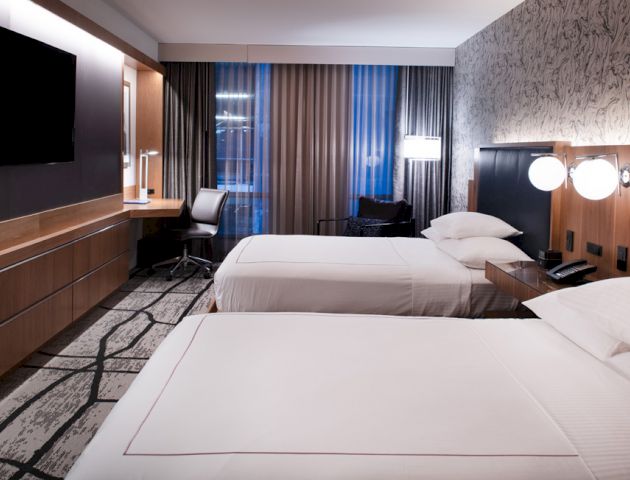 A modern hotel room featuring two double beds, a flat-screen TV, a desk with a chair, and stylish lighting and decor.