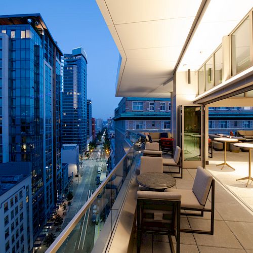 An outdoor terrace of a modern building with seating overlooks a city street lined with tall buildings at dusk, featuring a blue sky.