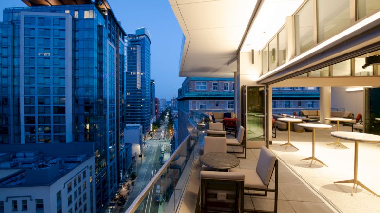 The image shows a modern urban balcony with seating overlooking a city street bordered by tall buildings during twilight.