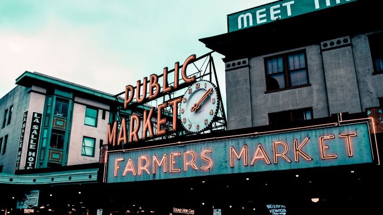 An outdoor market with neon signs reads "PUBLIC MARKET" and "FARMERS MARKET." The clock displays the time, and buildings are visible in the background.