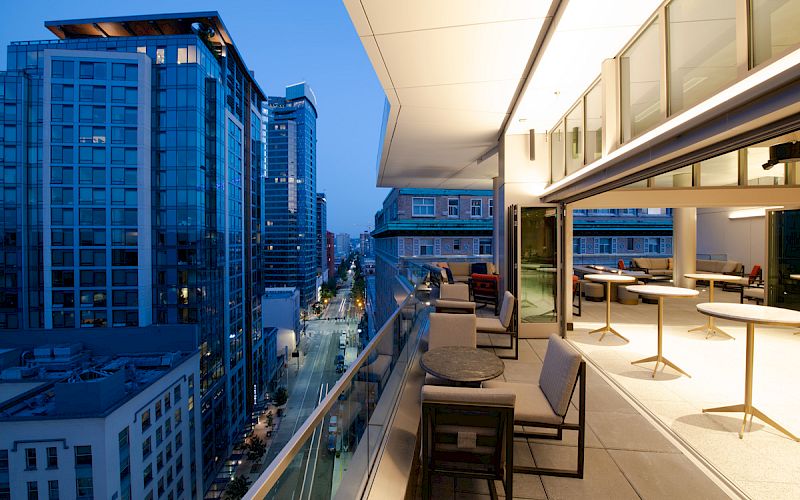 An urban view from a modern balcony with tables and chairs, overlooking a city street lined with tall buildings during early evening.