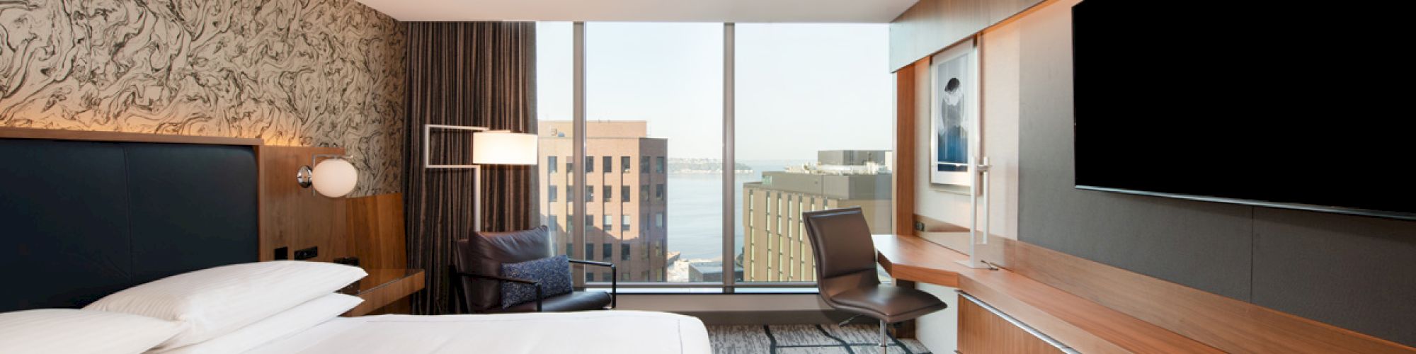 The image shows a modern hotel room with a large bed, a desk, and a chair, with a city view through large windows and a mounted TV.