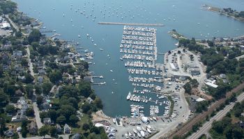 Aerial view of a coastal town featuring a marina with numerous docked boats, surrounded by residential and green areas, roads, and trees.