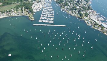 An aerial view of a marina with numerous boats docked and moored, surrounded by a coastal town and green landscape, with clear blue water.