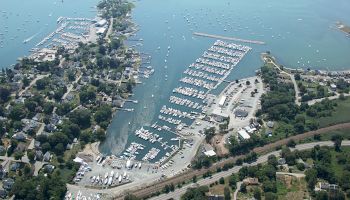 An aerial view of a coastal area featuring numerous boats docked at marinas, with surrounding buildings, roads, and greenery.