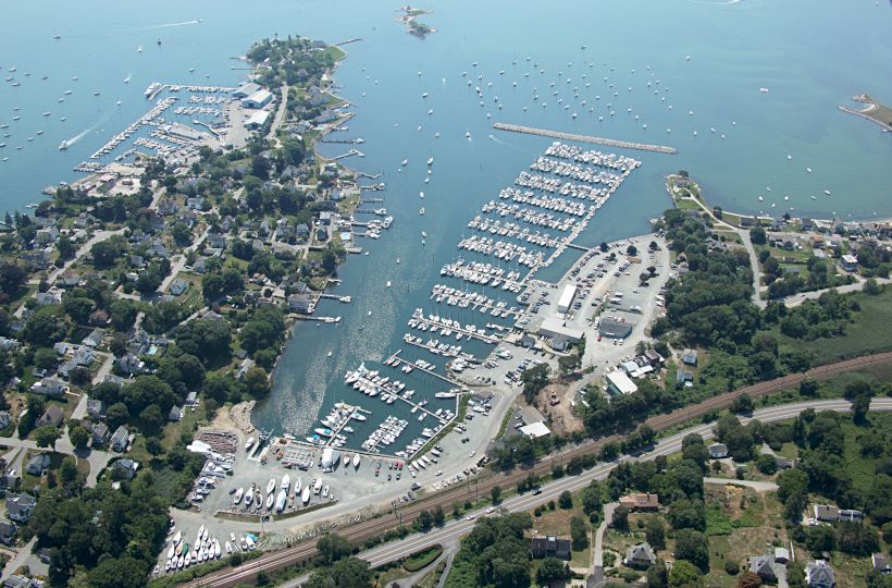 An aerial view of a coastal area featuring numerous boats docked at marinas, with surrounding buildings, roads, and greenery.