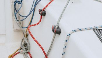 The image shows a section of a sailboat's deck with various ropes and lines, winches, and cleats. The background includes a dock.